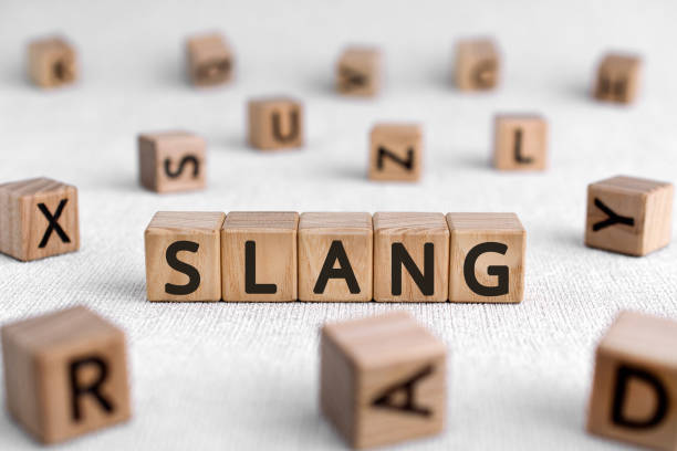 Wooden letter blocks spelling the word “SLANG” on a light fabric surface, surrounded by scattered alphabet blocks out of focus in the background.